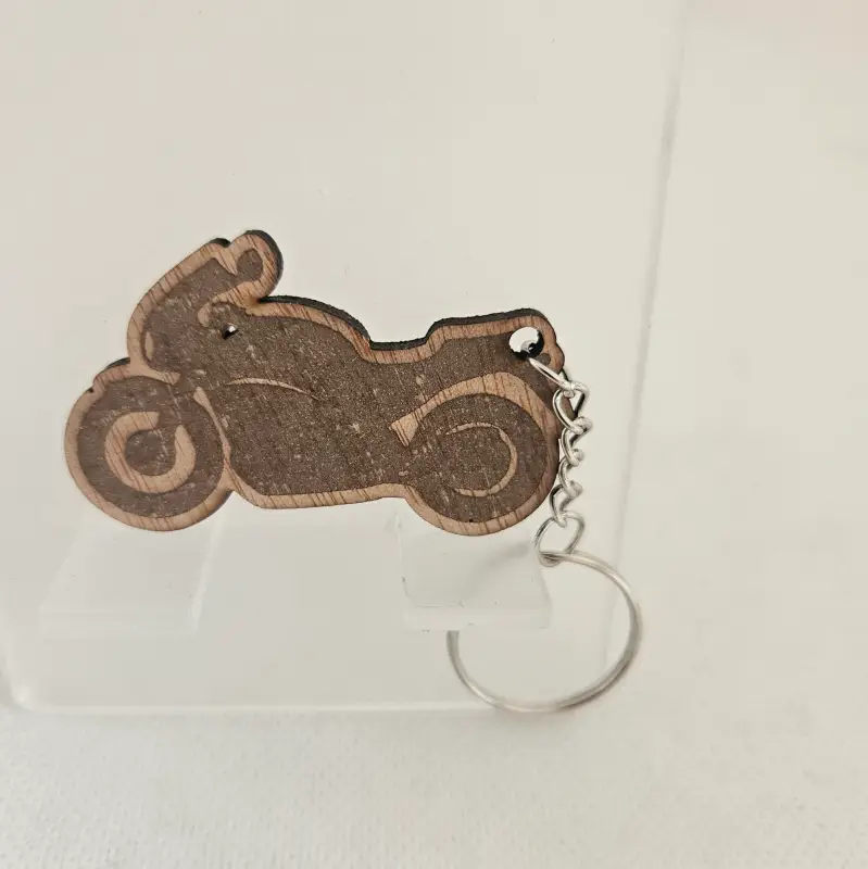 Porte clé en forme de moto découpé et gravé. il est attaché à un anneau métallique. Présenté sur un support et fond blanc.